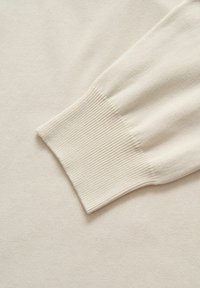 Licht beige gebreide trui detail met een ribbelmanchet en een gladde textuur, die de zachte stof en het eenvoudige ontwerp benadrukt.