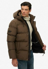 Mand iført en brun puffjakke og en cremefarvet turtleneck sweater, der holder jakken åben for at vise det sorte inderskud med logo tape.