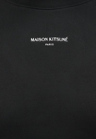 Μαύρο ύφασμα με λευκά κεντημένα γράμματα "MAISON KITSUNÉ" πάνω από μικρότερα γράμματα "PARIS", στο κέντρο κοντά στη λαιμόκοψη.