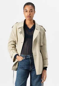Beige trench coat with shoulder epaulettes, buttoned front, and wide lapels, worn over a dark sweater and blue jeans. Smooth texture.