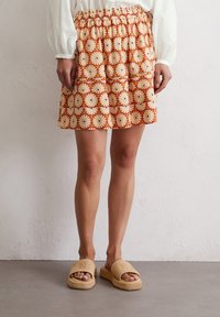 Gemusterter Rock mit elastischem Bund, der florale Designs in Orange, Creme und Dunkelblau zeigt. Kombiniert mit strukturierten beigen Riemchensandalen.