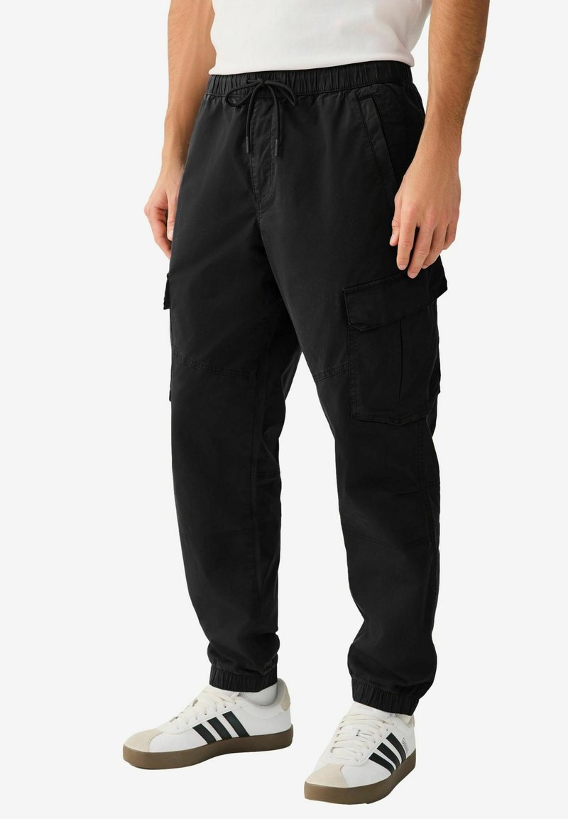 Pantalon cargo noir avec taille élastique et cordon de serrage, doté de poches latérales et d'ourlets resserrés. Porté avec des baskets blanches et grises.