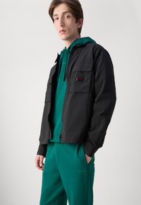 Svart lett zip-up jakke med to frontlommer, lagd over en grønn hettegenser og matchende grønne bukser, med HUGO-logoen.