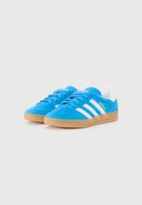 Tênis azul de camurça com três riscas brancas, sola de borracha natural e um calcanhar texturizado. Logos "Gazelle" e "Adidas" em dourado.