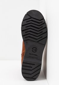 Bottine en daim marron avec une semelle en caoutchouc présentant un motif de bande de roulement en zigzag. Logo imprimé sur la semelle. Bottine appuyée contre une surface blanche.