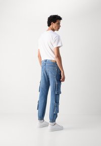 Pantalones cargo de mezclilla azul claro con múltiples bolsillos laterales, puños elásticos y una pequeña etiqueta en el bolsillo trasero. Combinados con una camiseta blanca.