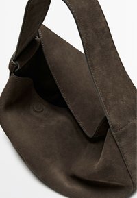Brown suede handbag with a soft texture, folded flap closure, and a curved strap. Features a subtle embossed circle detail.