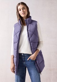 Lila gepolsterte Weste mit hohem Kragen, mit diagonaler Steppung, kombiniert mit einem hellen Pullover und blauen Jeans. Mit Reißverschluss und Seitentaschen.