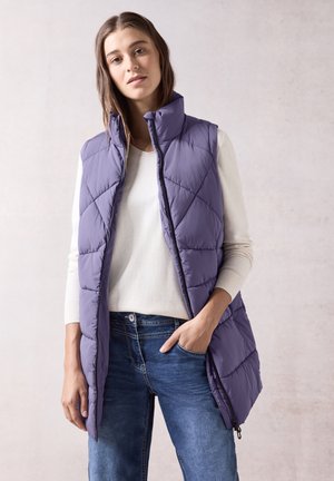 Gilet rembourré violet avec un col montant, présentant un matelassage diagonal, associé à un pull léger et un jean bleu. Fermeture éclair et poches latérales.