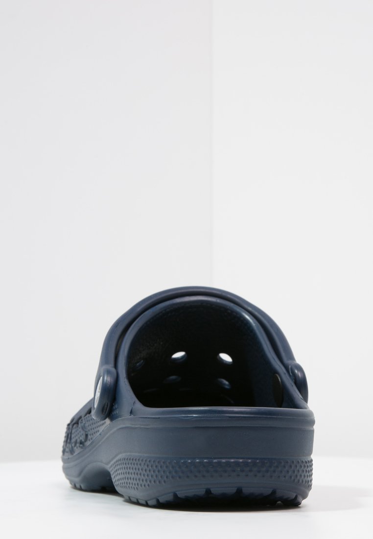 Zoccolo in gomma blu scuro con fori di ventilazione, cinturino posteriore pronunciato, suola texturizzata e superficie liscia. La vista posteriore mostra un design arrotondato.