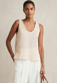 Strick-Tanktop in hellbeige mit weitem Rundhalsausschnitt, strukturiertem Stoff und seitlichen Schlitzen, kombiniert mit leichten weißen Hosen.