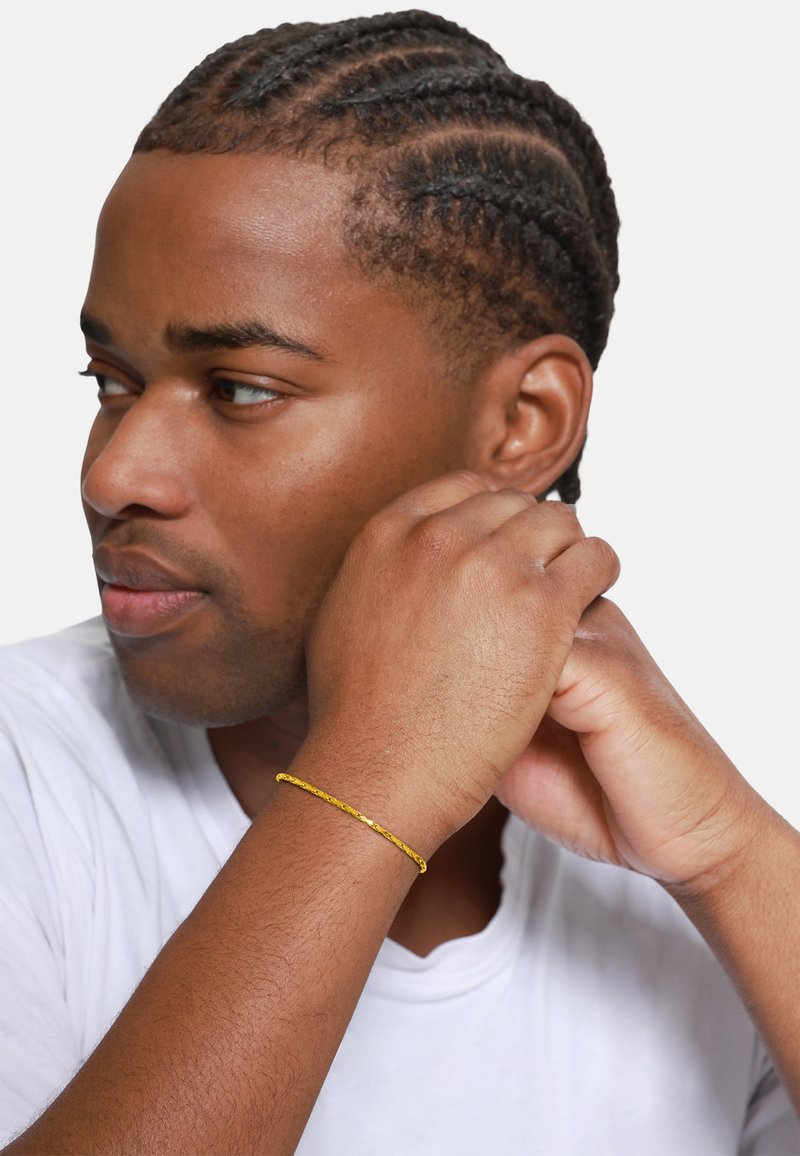 Gouden armband met complexe textuur, gedragen om de pols, gecombineerd met een eenvoudig wit T-shirt. Haar in nette cornrows gestyled.