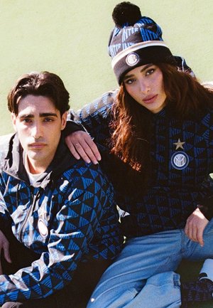 Man en vrouw zitten op het gras en dragen bijpassende blauwe jasjes met een patroon van Inter Milan; de vrouw draagt ook een Inter Milan muts en lichte spijkerbroek.