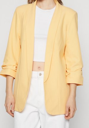 Gul kavaj med en något strukturerad design, som har en uppskuren krage och uppvikt ärmar, i kombination med en vit crop top och vita byxor.