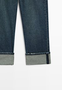 Ciemnoniebieskie dżinsy z lekkim przetarciem u dołu. Nogawki podwinięte, odsłaniając jasnoszary materiał od spodu. Widoczne standardowe przeszycia.