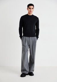 Pull en maille gris foncé, pantalon à rayures lâche, et chaussures noires épaisses. Le modèle se tient avec les mains dans les poches devant un fond blanc.