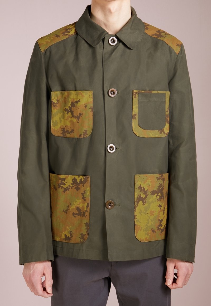 Veste verte avec des patches en camouflage sur les épaules et les poches. Dotée d'une fermeture à boutons, d'un col classique et de plusieurs poches avant.