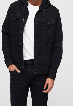 Mann trägt schwarze Knopfleiste-Jacke mit Kapuze über weißem T-Shirt und schwarzer Hose, eine Hand in der Tasche, vor einfachem Hintergrund.