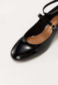 Chaussure Mary Jane en cuir verni noir avec bout arrondi, bride à la cheville et semelle intérieure en cuir beige, vue en angle par le dessus.