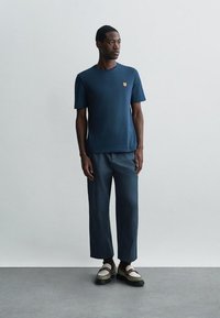 Hombre de pie, vistiendo una camisa de manga corta azul marino con un parche de tigre, pantalones a juego, calcetines oscuros y mocasines bicolor.