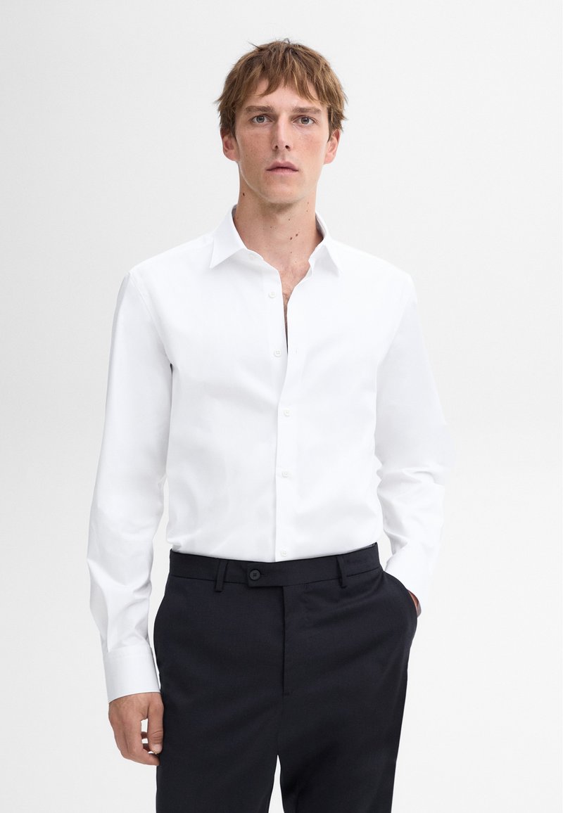 Weißes Langarmhemd mit Button-Down-Kragen und Knopfleiste, getragen mit dunklen, taillierten Hosen. Glattes Material, sauberes Design.