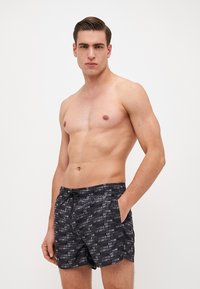 Fekete úszóshort, fehér színű ismétlődő logó mintával. Az anyag könnyű, rugalmas derékkal és oldalsó zsebekkel rendelkezik.