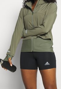 Olivgrön zip-up-jacka med långa ärmar och reflekterande detaljer, matchad med svarta träningsshorts med en vit Adidas-logga.