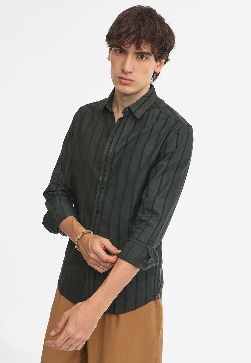 Camisa de botones a rayas verde oscuro con cuello, mangas largas y puños enrollados. Textura suave con un ligero brillo. Llevada con pantalones beige.