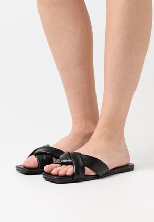 Stopy w czarnych, płaskich sandałach z szerokimi, miękkimi paskami skrzyżowanymi na białym tle.
