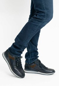 Černé a hnědé sportovní boty s texturovanou kůží a semišovými akcenty, s šněrovacím designem a gumovou podrážkou, nošené s tmavými džíny.