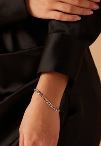 Zilveren kettingarmband om een pols, met onderling verbonden schakels. De hand rust op een achtergrond van zwart satijn.