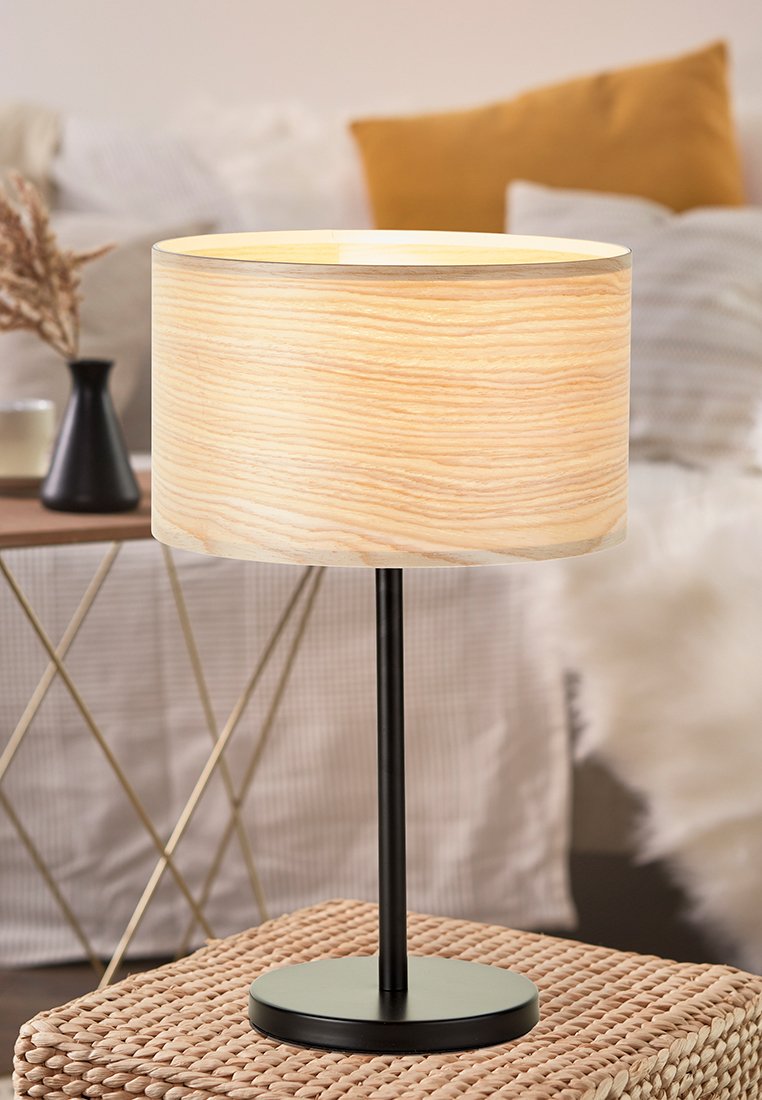 Stolní lampa s cylindrickým stínidlem ze světlého dřeva a černým kovovým podstavcem stojící na pleteném ottomanu v útulném obývacím pokoji.