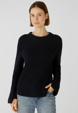 Marineblauwe gebreide trui met lange mouwen, ribbeltextuur, ronde hals en zijslitsen, gecombineerd met versleten blauwe jeans.