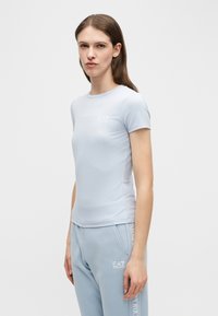 Γυναίκα με μακριά καστανά μαλλιά που φοράει ανοιχτό μπλε μπλουζάκι EA7 Emporio Armani και αντίστοιχο παντελόνι φόρμας, στέκεται μπροστά σε ένα απλό ανοιχτό φόντο.