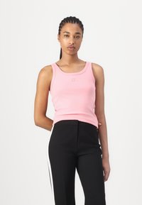 Débardeur rose clair avec un col rond, arborant un petit logo en relief, associé à un pantalon noir avec des bandes blanches sur les côtés.