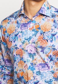 Camisa de botones floral con un fondo azul claro. El patrón incluye flores naranjas, moradas y azules con hojas verdes. Material de algodón.