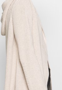 Beige pletený svetr s rolákem, nabízející volný střih a jemnou texturu. Design obsahuje otevřené strany a měkký, útulný vzhled.