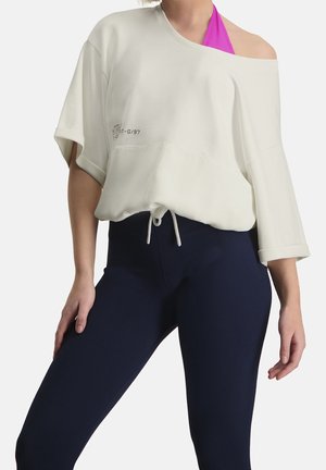 Person trägt einen off-white asymmetrischen, locker sitzenden Oberteil über einem pinkfarbenen Badeanzug-Träger mit marineblauen, eng anliegenden Leggings vor weißem Hintergrund.