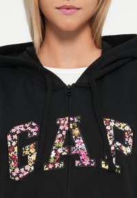 Felpa nera con lettere 'GAP' decorate da un motivo floreale. Il materiale sembra morbido; presenta una cerniera completa e cappuccio, con una finitura in tessuto testurizzata.