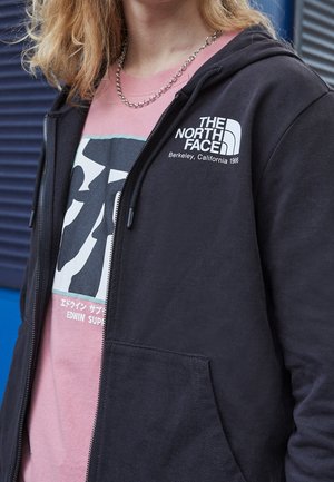 Osoba nosząca czarną bluzę z kapturem na zamek z logo "The North Face" i różowy t-shirt z grafiką pod spodem, z jasnobrązowymi włosami i łańcuszkiem na szyi.