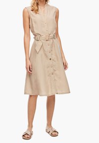 Beige, ärmlös klänning med V-ringning, framknappar och ett midjebälte. Tyget har en slät textur och klänningen har en utställd kjol.
