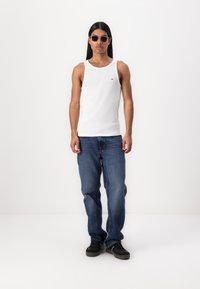 Weißes geripptes Tanktop, blaue Jeans und schwarze Sneakers, getragen von einer Person mit langen dunklen Haaren und runden Sonnenbrillen, die vor einem weißen Hintergrund steht.