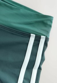 Zielone sportowe szorty o gładkiej fakturze, z dwoma białymi pionowymi paskami i paskiem w pasie w ciemniejszym odcieniu zieleni.