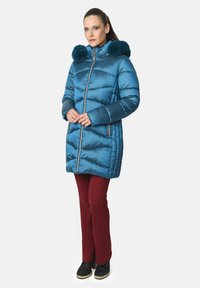 Chaqueta acolchada en color teal con capucha forrada de piel, cierre de cremallera al frente y bolsillos laterales. Combinada con pantalones burdeos ajustados y botas negras.