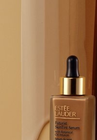 Sérum teint futuriste d'Estée Lauder dans un flacon en verre avec un capuchon doré et un compte-gouttes noir, contenant un liquide beige lisse à l'intérieur.