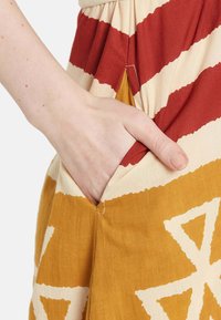 Baumwollkleid mit kräftigen roten und senfgelben Streifen, ausgestattet mit einer Seitentasche. Texturierter Stoff mit einem geometrischen Muster in Senfgelb.