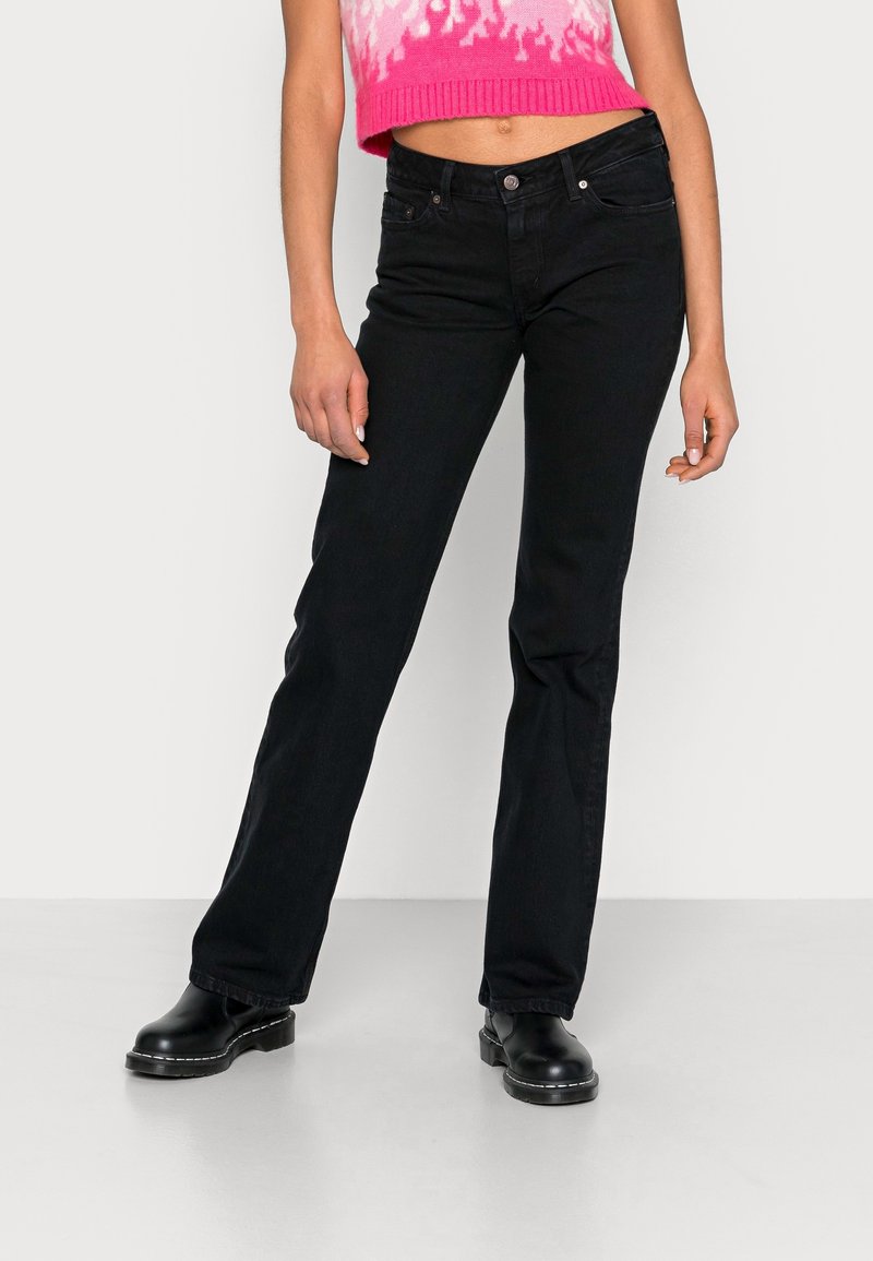 Femme portant un jean noir droit, des bottes noires et un pull court rose et blanc à motifs, debout devant un fond uni.