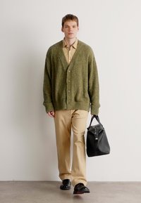 Groene, gestructureerde cardigan met een v-hals, knopen en lange mouwen, gecombineerd met beige broeken. Draagt een zwarte tas en zwarte schoenen.