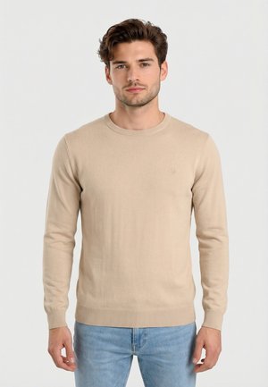 Joven con cabello corto y castaño, vestido con un suéter beige de manga larga y jeans azul claro, posando frente a un fondo blanco liso.