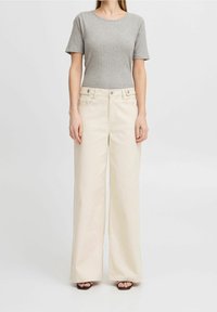 Grå ribbet kortærmet top kombineret med cremefarvede bukser med vide ben, der har forlommer og bæltestropper, placeret mod en ensfarvet baggrund.